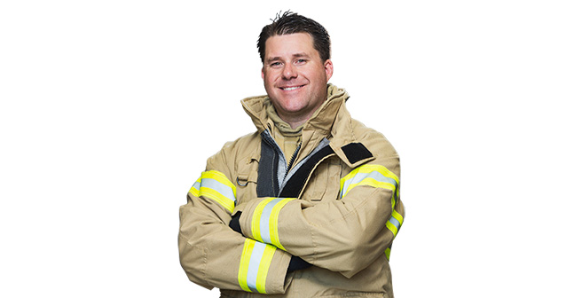 A smiling male firefighter wearing beige and partly high-visibility PPE carries with arms crossed
